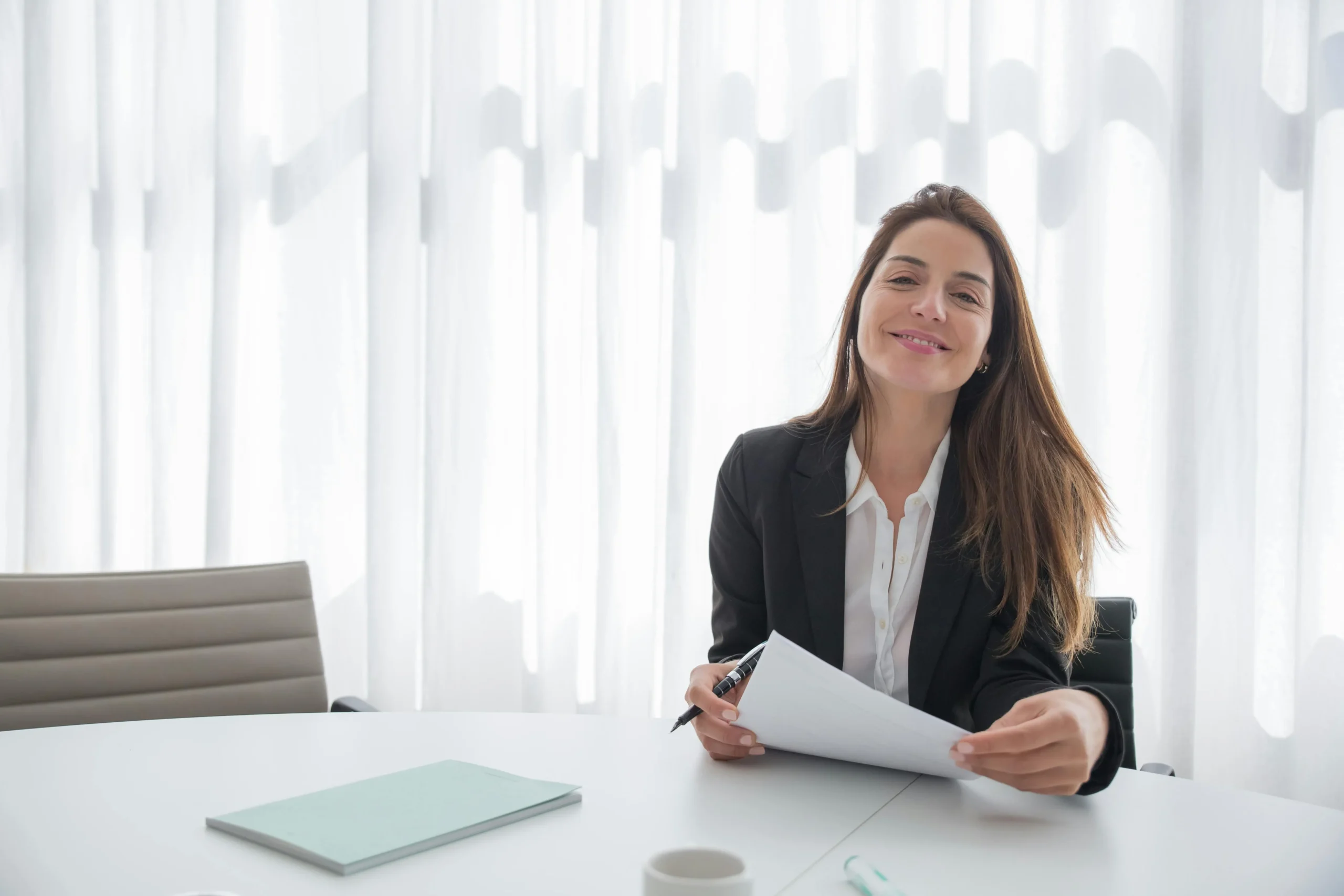 Lächelnde Beraterin im Business-Outfit sitzt mit Dokumenten und Stift an einem weißen Besprechungstisch.